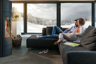Des personnes sont détendues sur un canapé sombre et moderne devant un feu de cheminée crépitant, avec vue sur un paysage enneigé à travers de grandes baies vitrées panoramiques. Une corbeille remplie de bois se trouve à côté de la cheminée, des magazines et des chaussures se trouvent dans le salon.