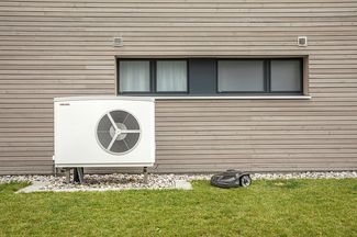 Vue extérieure d’une maison moderne avec façade en bois; devant le mur se trouve une pompe à chaleur air/eau, à côté d’un robot-tondeuse autonome sur la pelouse.